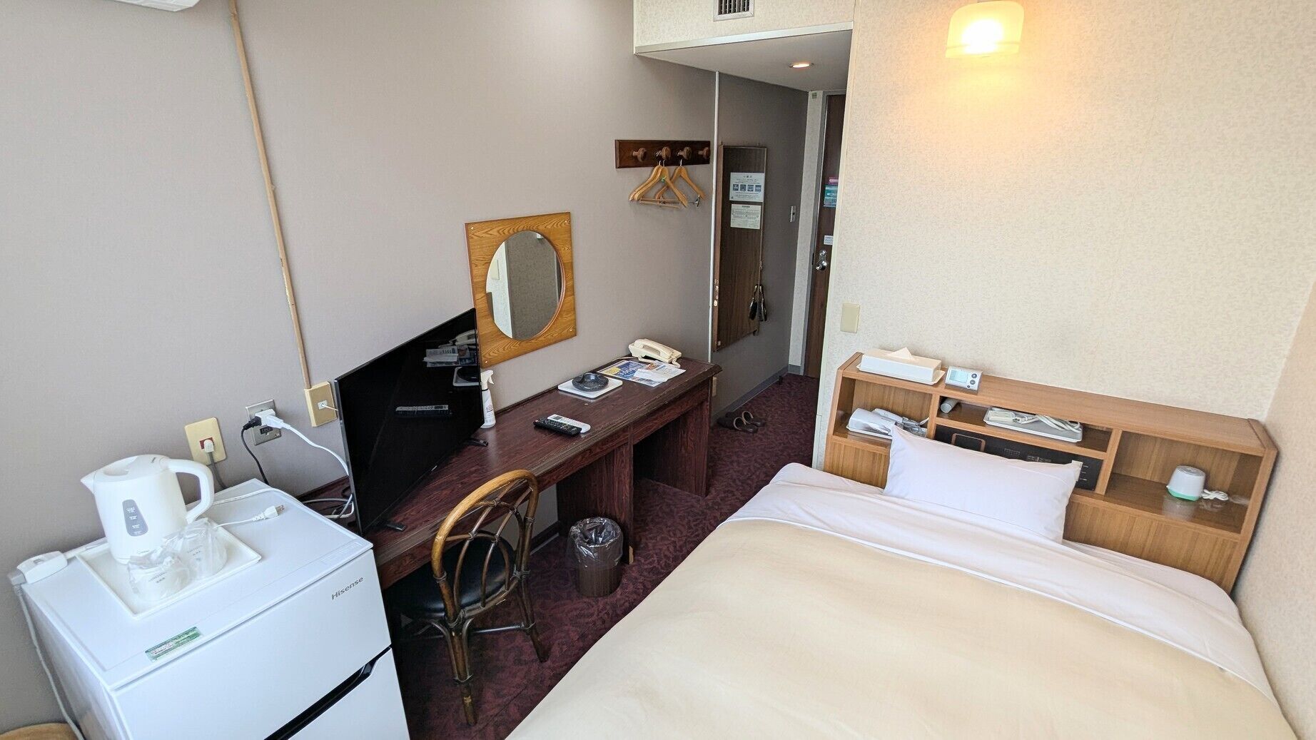 Habitación de hotel con cama, escritorio con silla, espejo, teléfono, estante pequeño con artículos de aseo y un cesto de basura.
