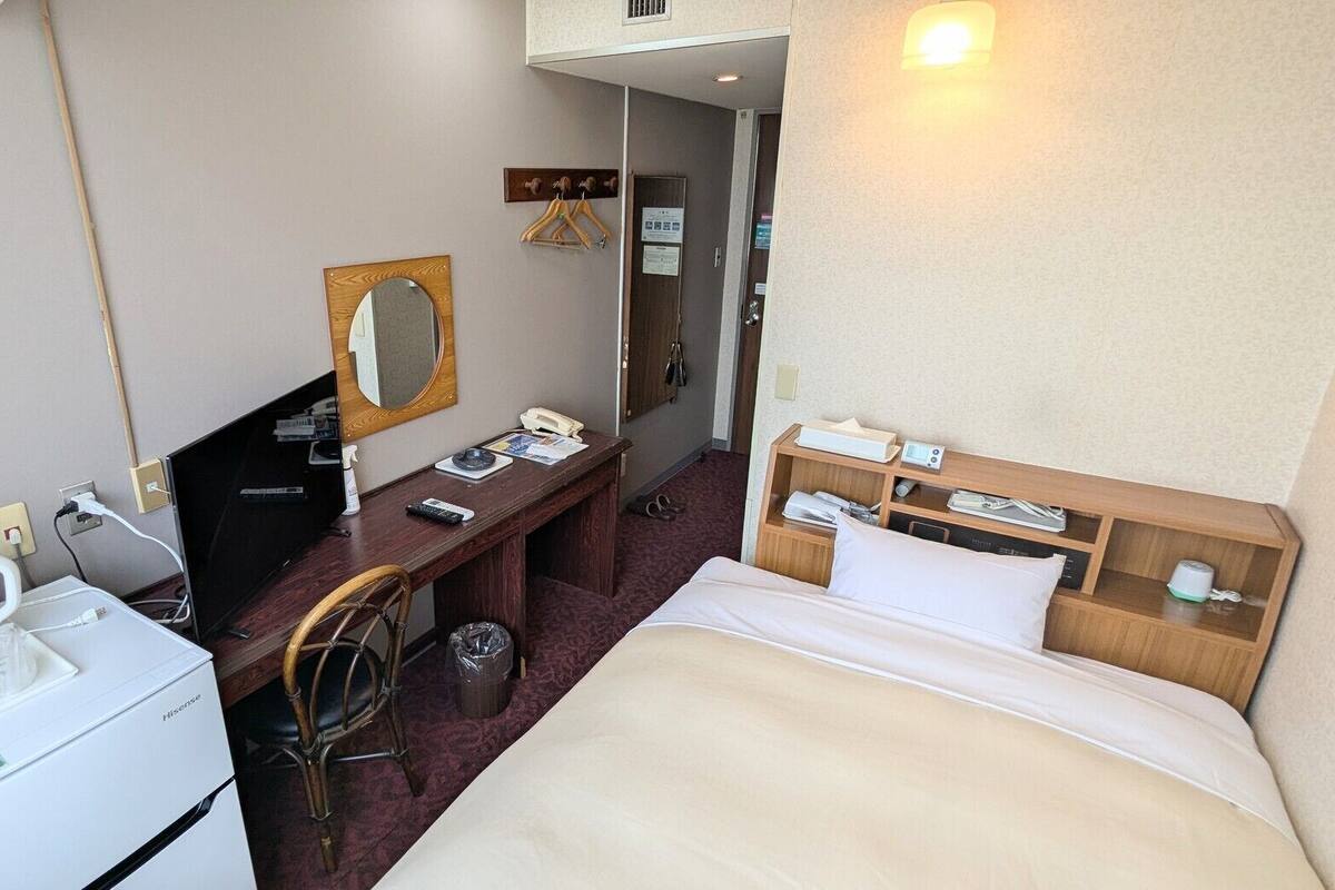 Habitación de hotel con cama, escritorio con silla, espejo, teléfono, estante pequeño con artículos de aseo y un cesto de basura.