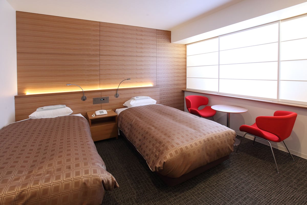 Habitación de hotel con dos camas, un cabecero de madera, una mesita de noche pequeña, una silla roja, una mesa redonda y un ventanal grande.
