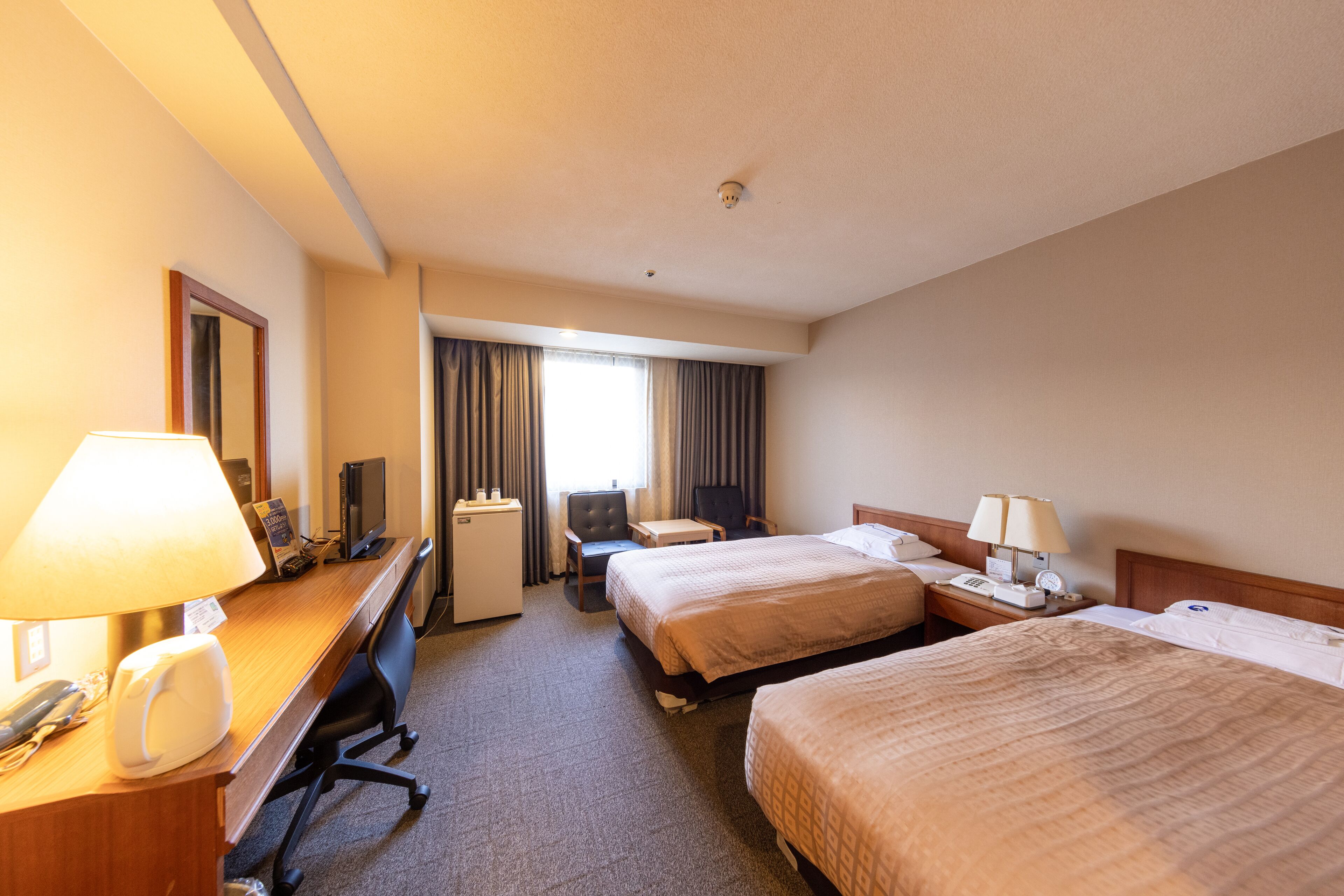 A hotel room with two beds, a desk with a computer, a chair, a desk lamp, and a telephone.