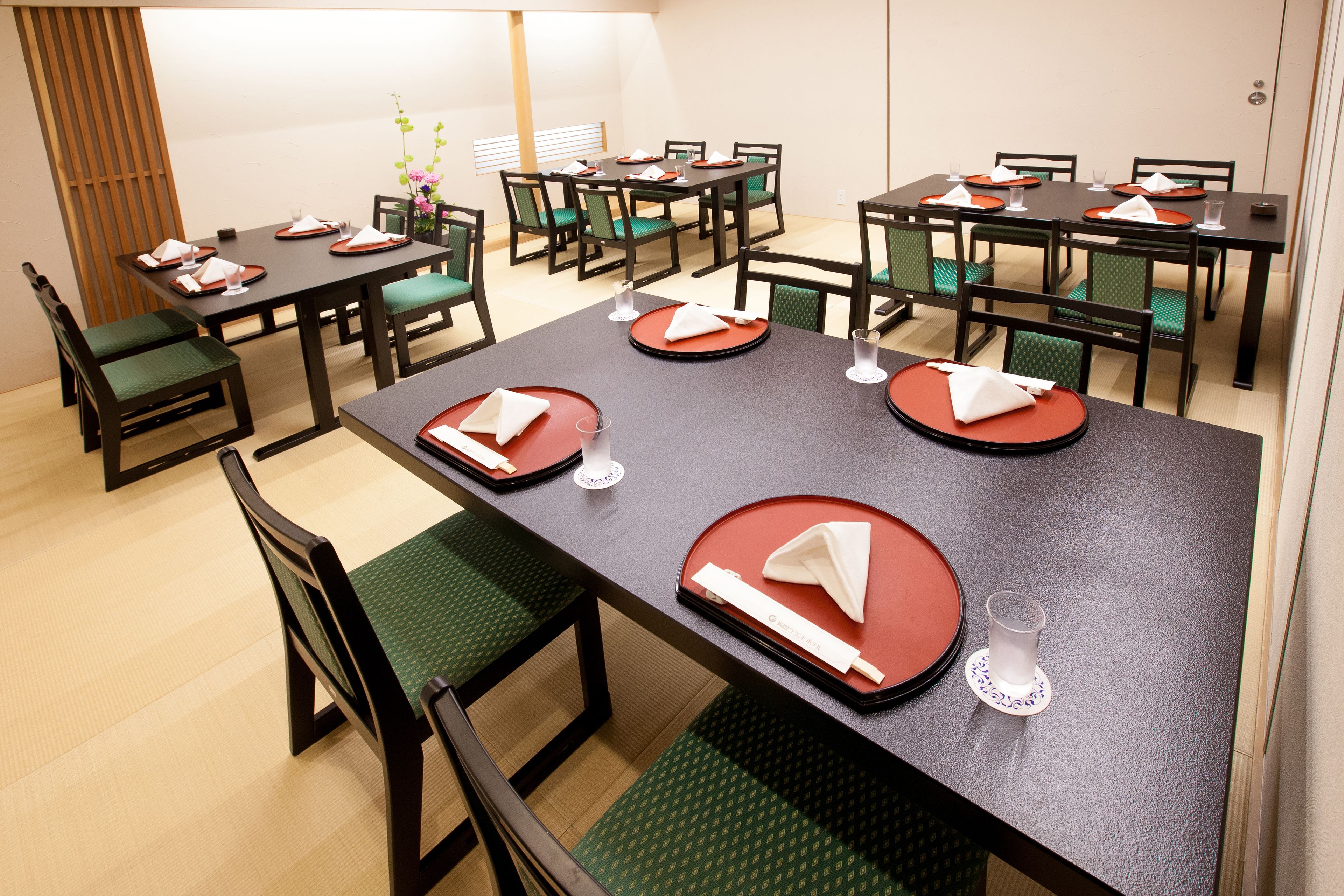 Comedor con varias mesas dispuestas, manteles individuales rojos, servilletas blancas y sillas con cojines verdes.