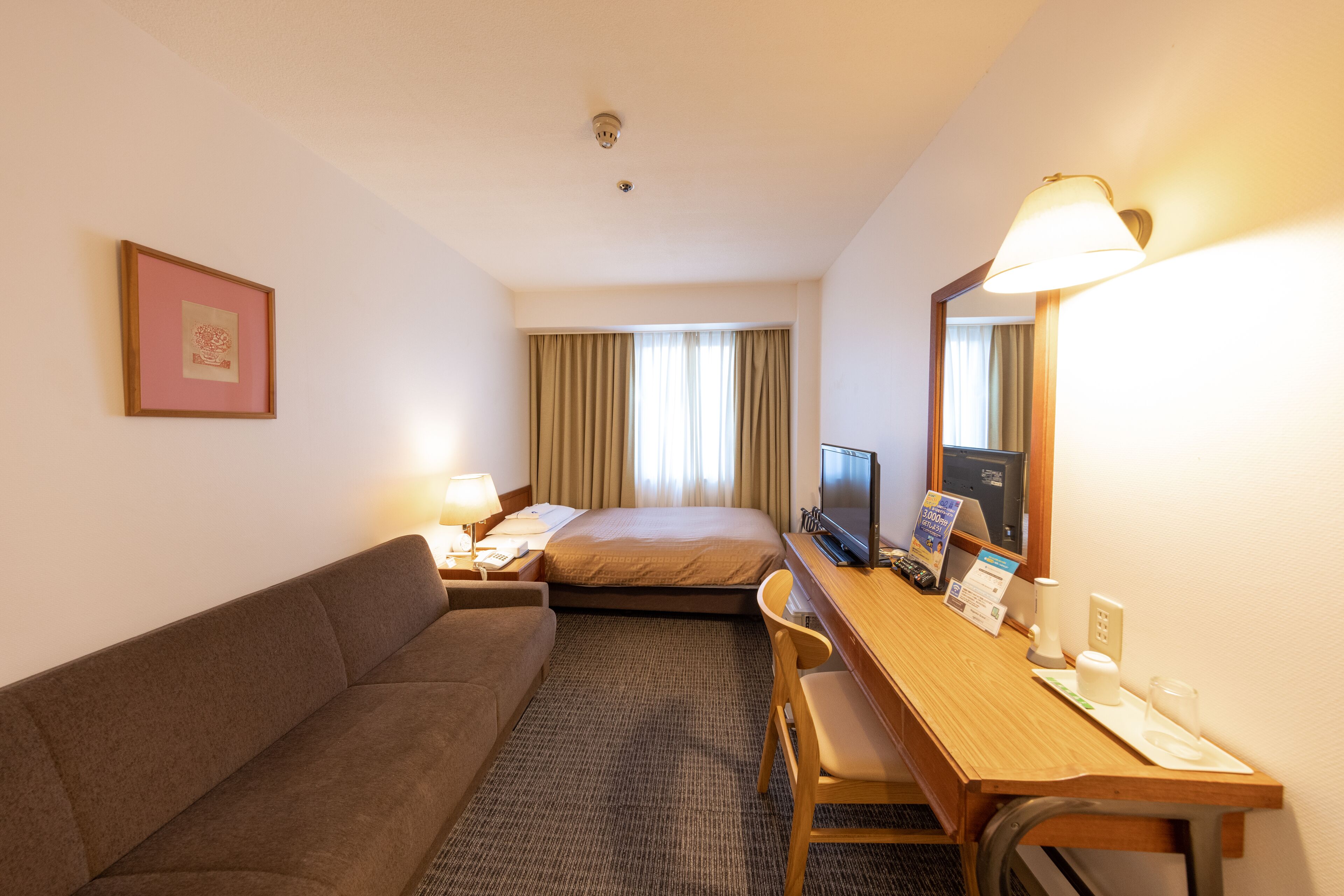 A hotel room with a bed, a desk with a computer, a chair, a sofa, a lamp, and a framed picture on the wall.