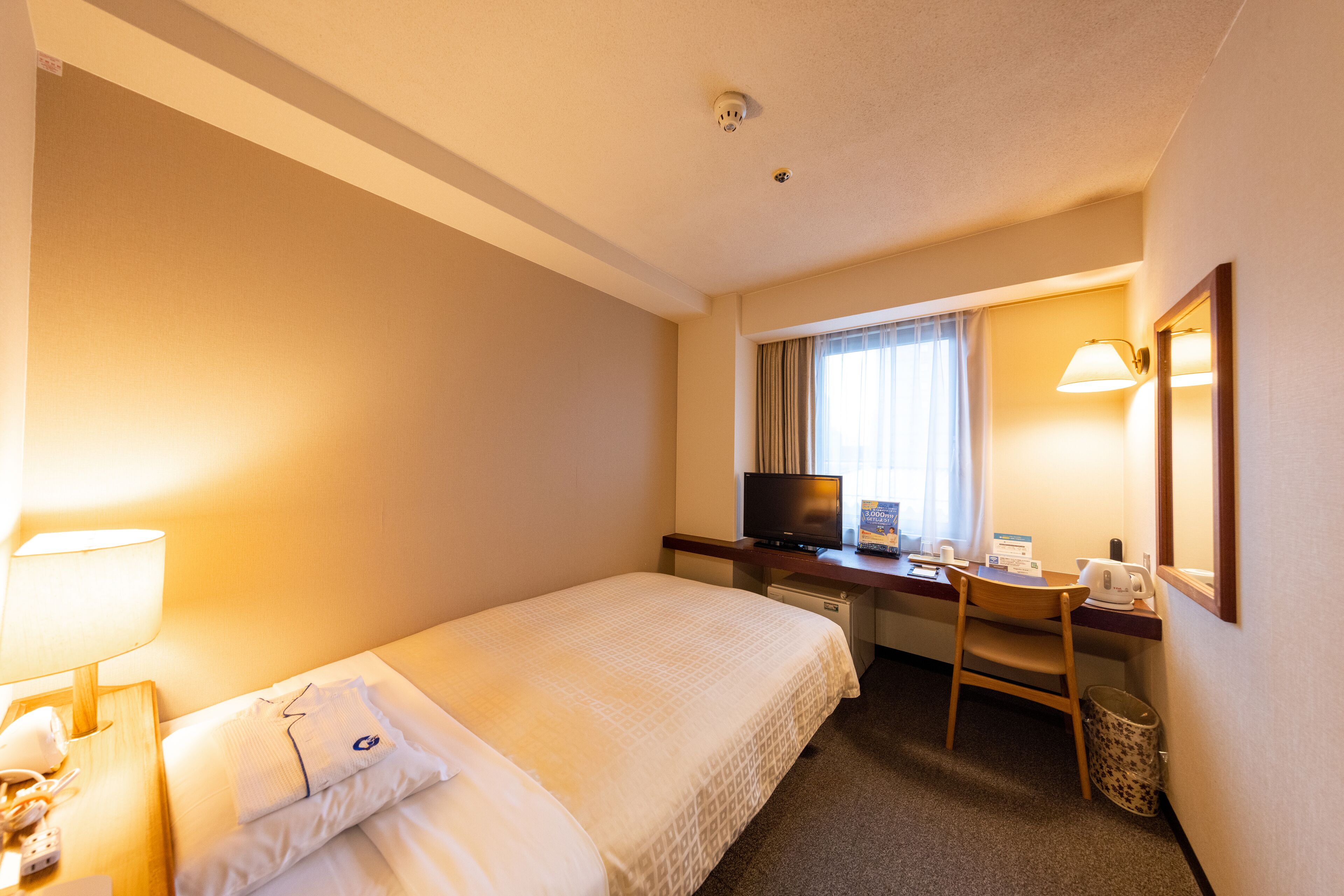 Habitación de hotel con cama, escritorio, silla, lámpara, televisión y ventana con cortinas.