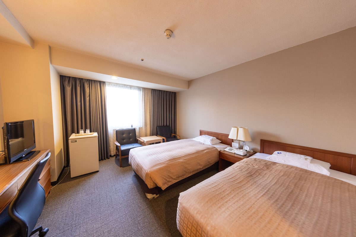 Una habitación de hotel con dos camas, un escritorio con computadora, un refrigerador pequeño y un televisor.