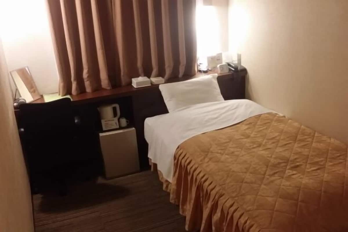 A hotel room with a bed, a desk with a lamp, a small table with a coffee maker, and curtains.