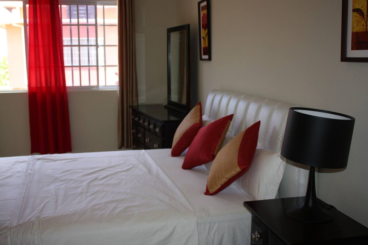 A neatly made bed with white linens and red and beige pillows, a black bedside lamp, a dresser with a mirror, and a window with red curtains.