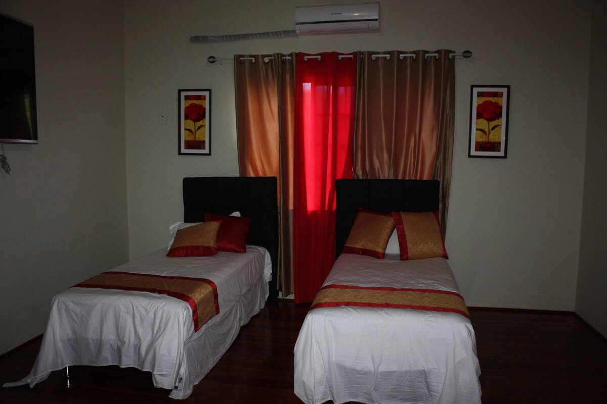 A hotel room with two single beds, each with a red and gold bedspread, a red curtain, and a wall-mounted air conditioner.
