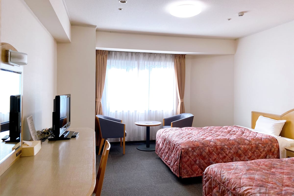 Quarto de hotel com duas camas, uma escrivaninha com computador e televisão, uma mesinha e uma janela com cortinas.
