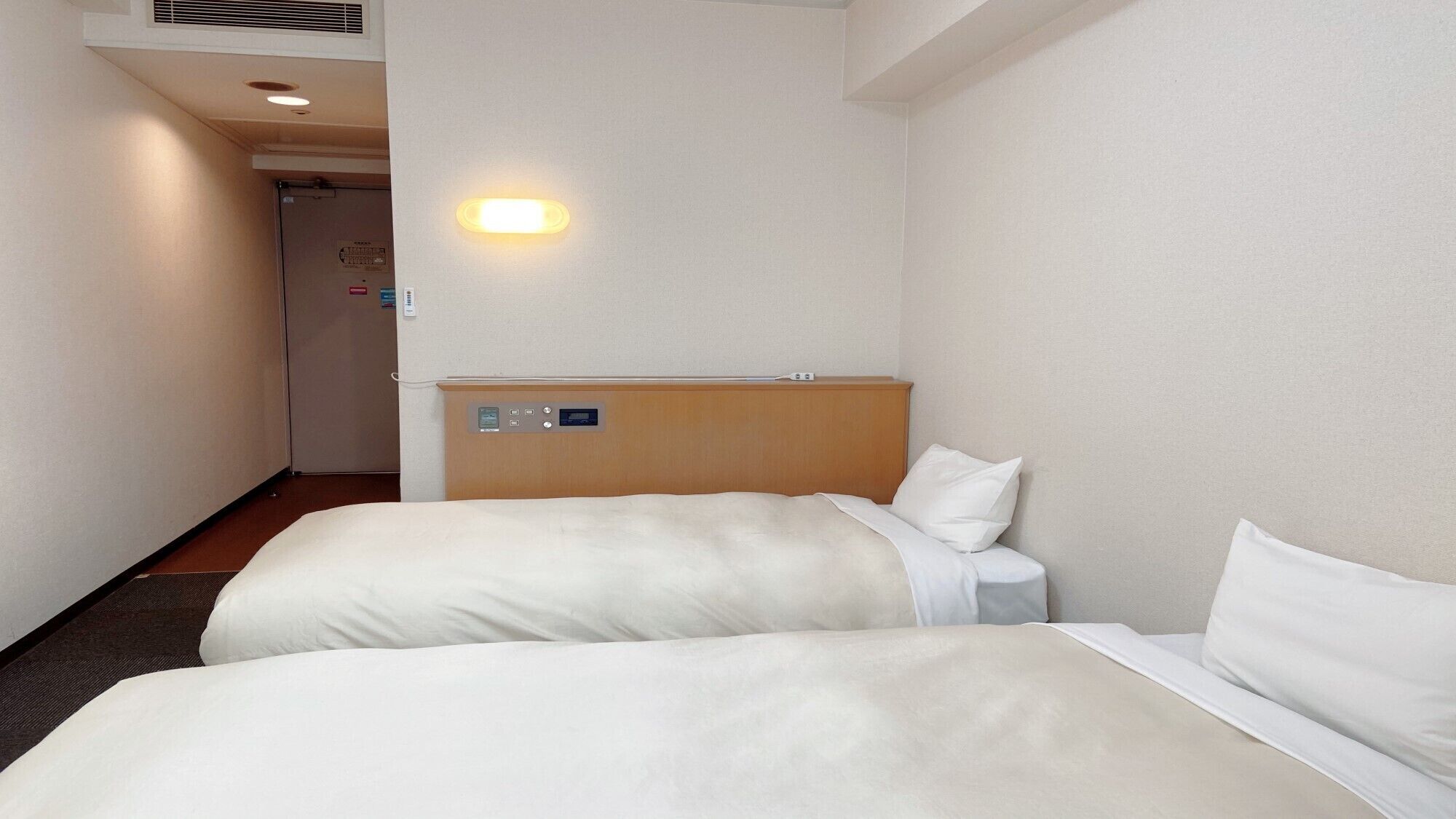 A hotel room with two beds, a wooden headboard, a bedside table, and a wall-mounted control panel.