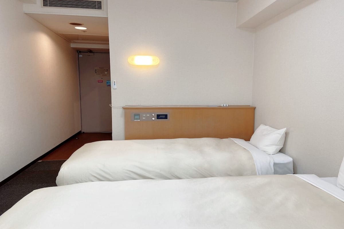 Quarto de hotel com duas camas, cabeceira de madeira, mesa de cabeceira e painel de controle fixado na parede.