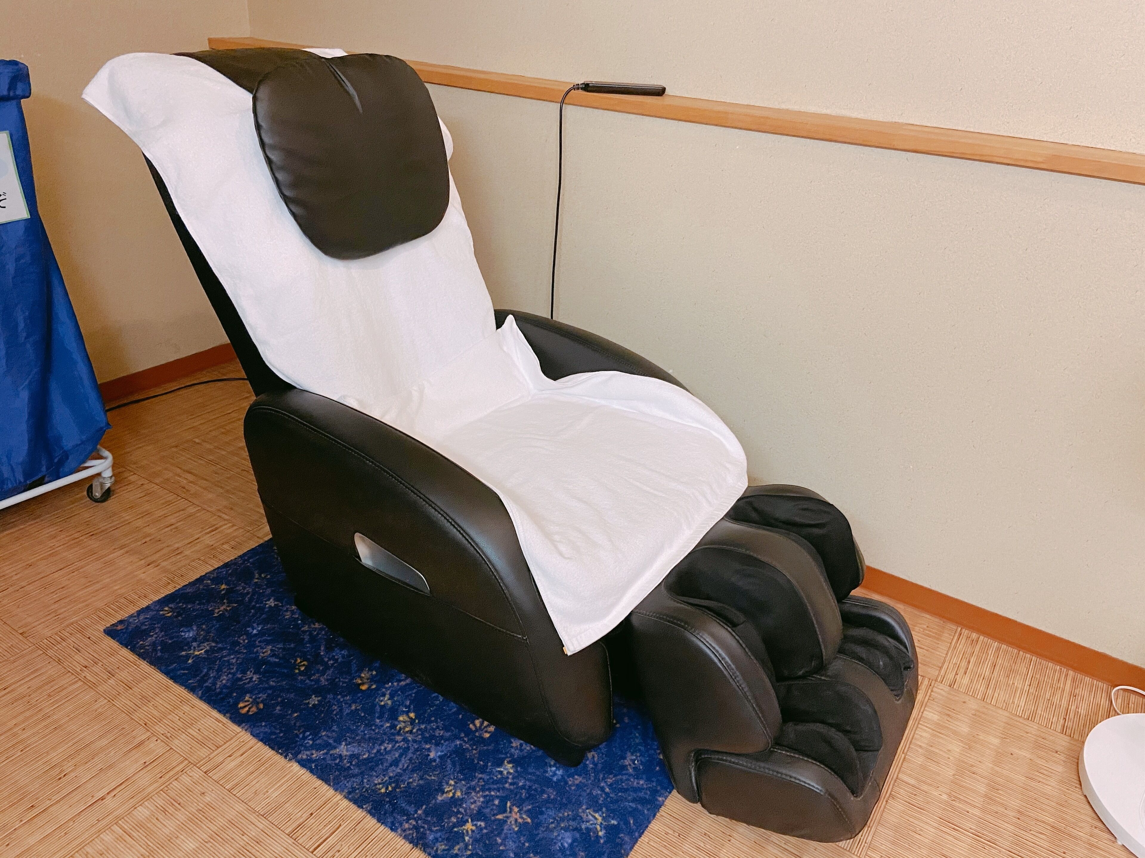 A massage chair with a footrest, a blue patterned rug, and a wooden shelf against the wall.