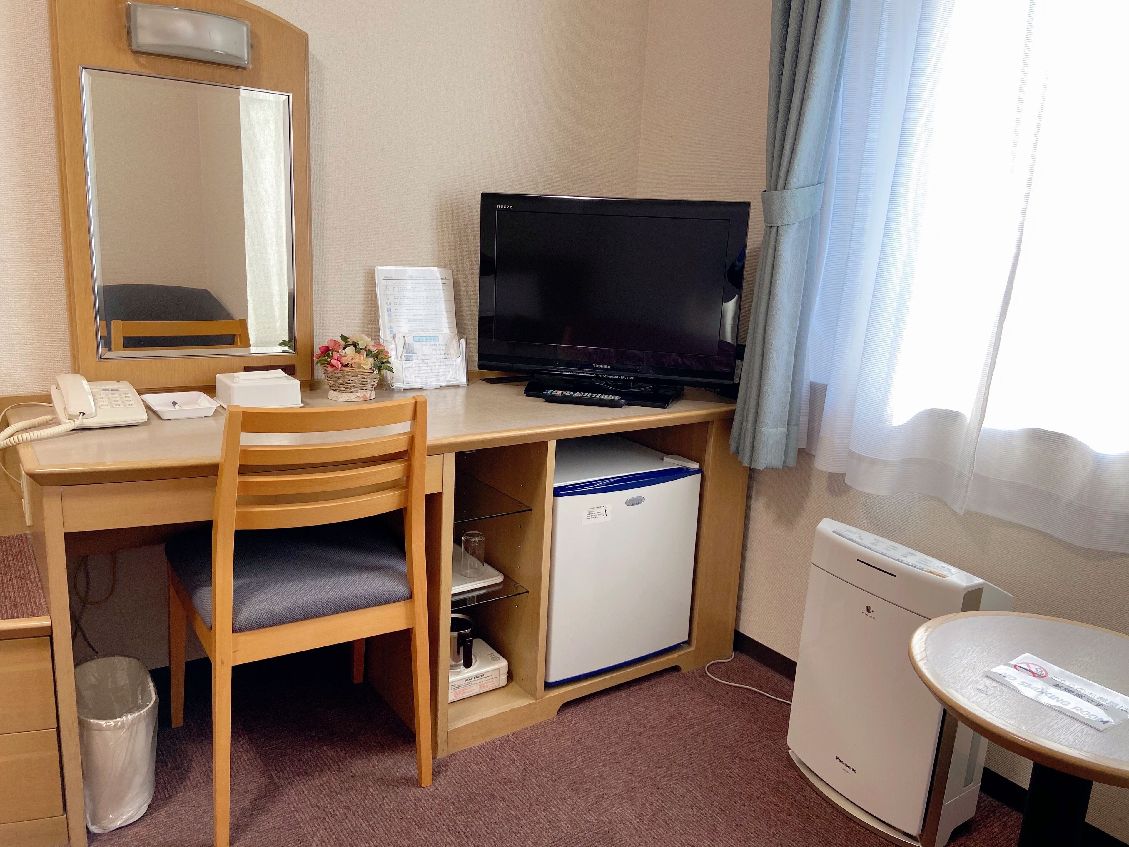 A hotel room with a wooden desk, a television, a small refrigerator, a trash bin, and a bedside table with a sign.