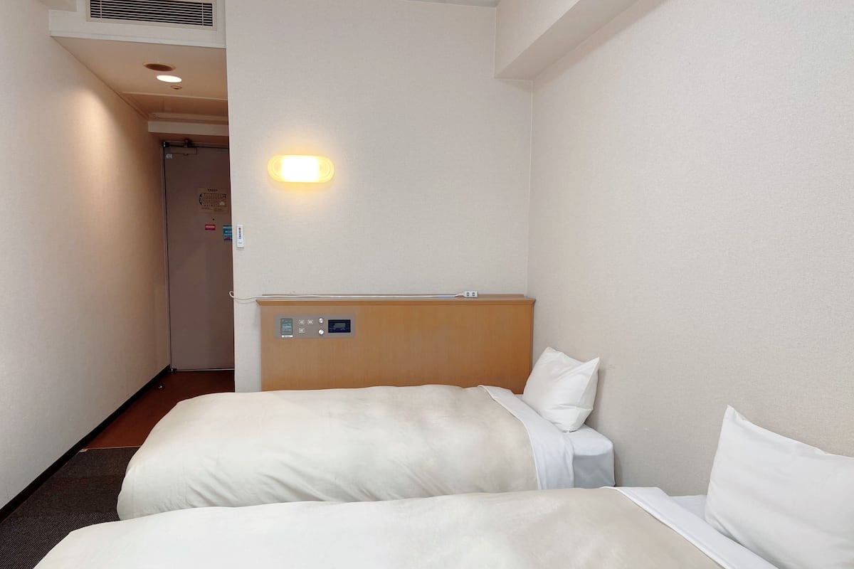 Quarto de hotel com duas camas, uma cabeceira de madeira, uma mesinha de cabeceira e uma luminária fixada na parede.