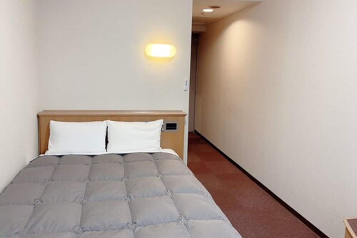 Quarto de hotel com cama, mesa de cabeceira, luminária na parede e porta para outro cômodo.