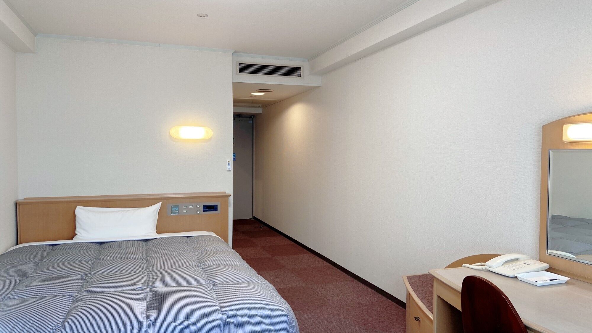 A hotel room with a bed, bedside controls, a desk with a phone, and a mirror.