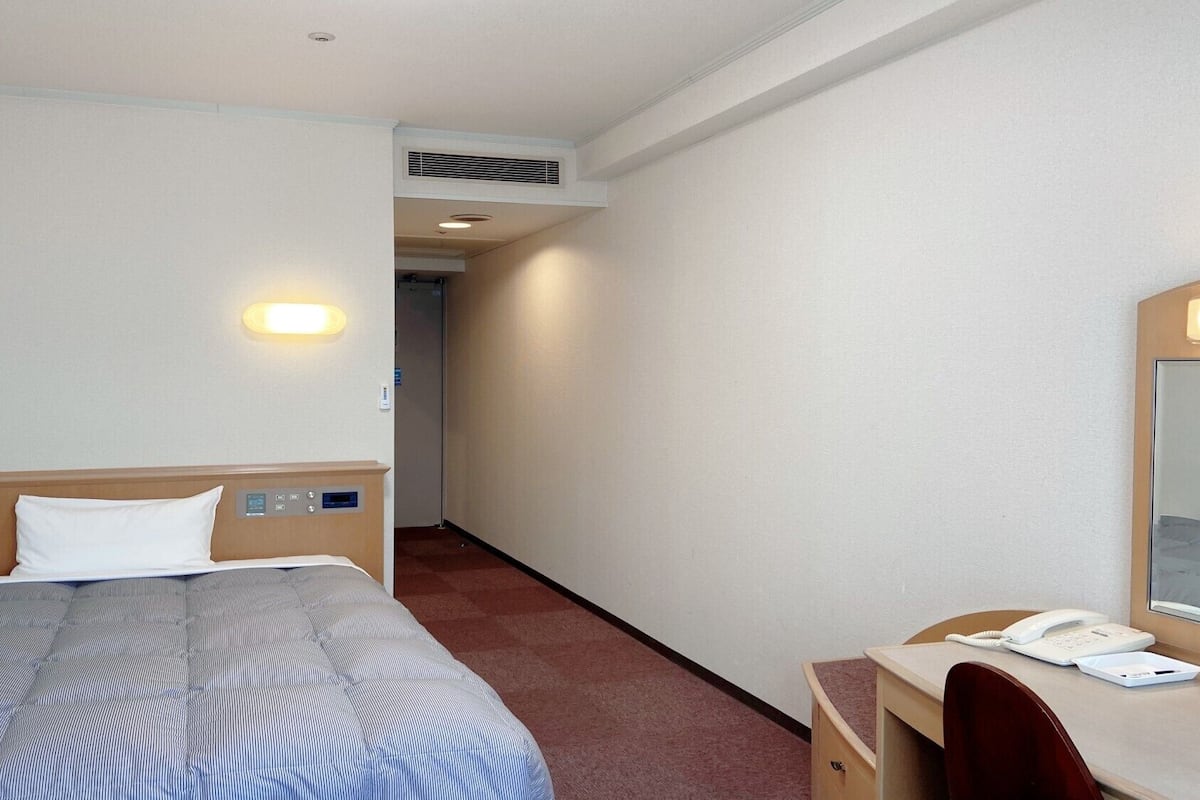Quarto de hotel com cama, controles laterais, uma escrivaninha com telefone e um espelho.