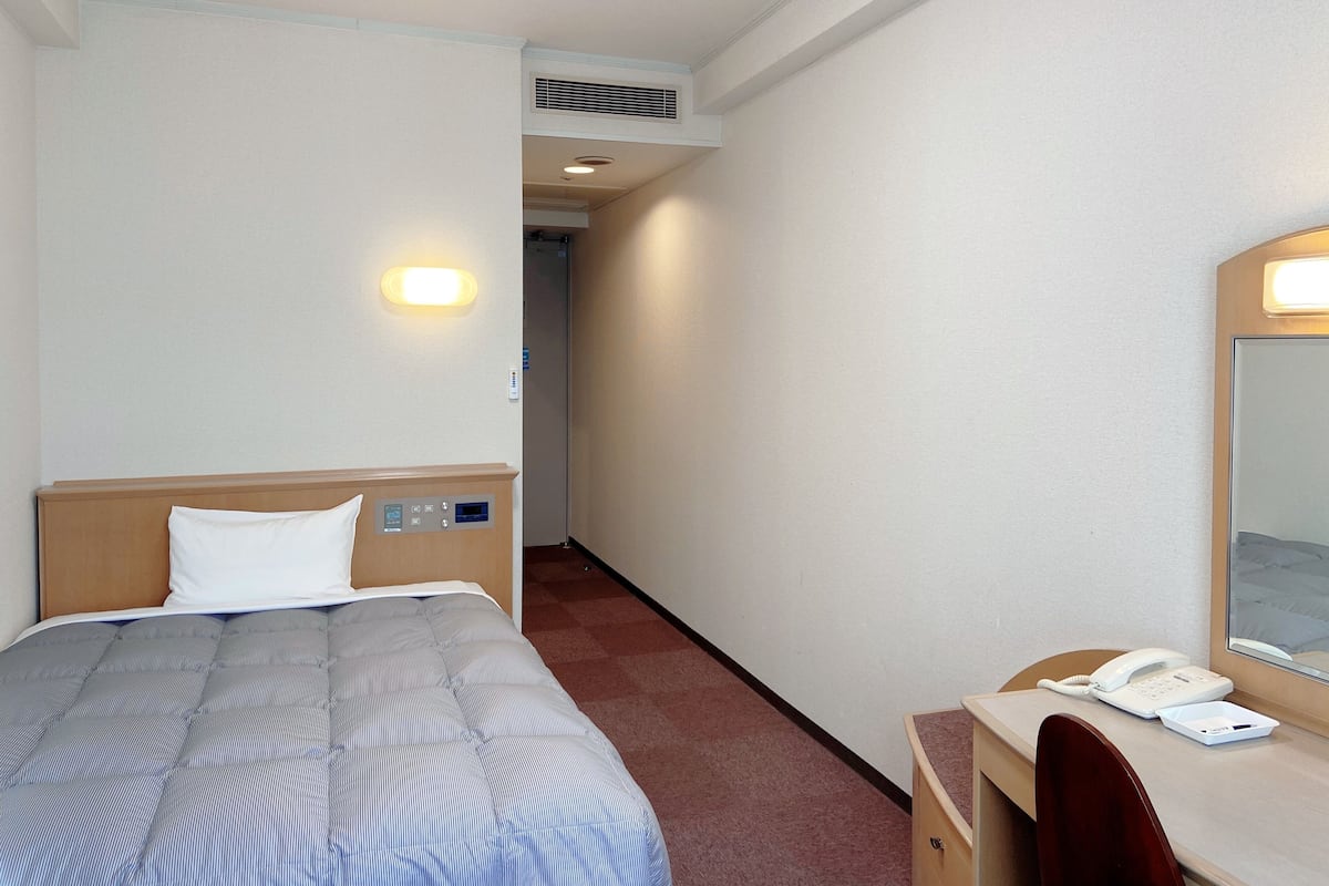 Quarto de hotel com cama, controles ao lado da cama, escrivaninha com telefone e espelho.