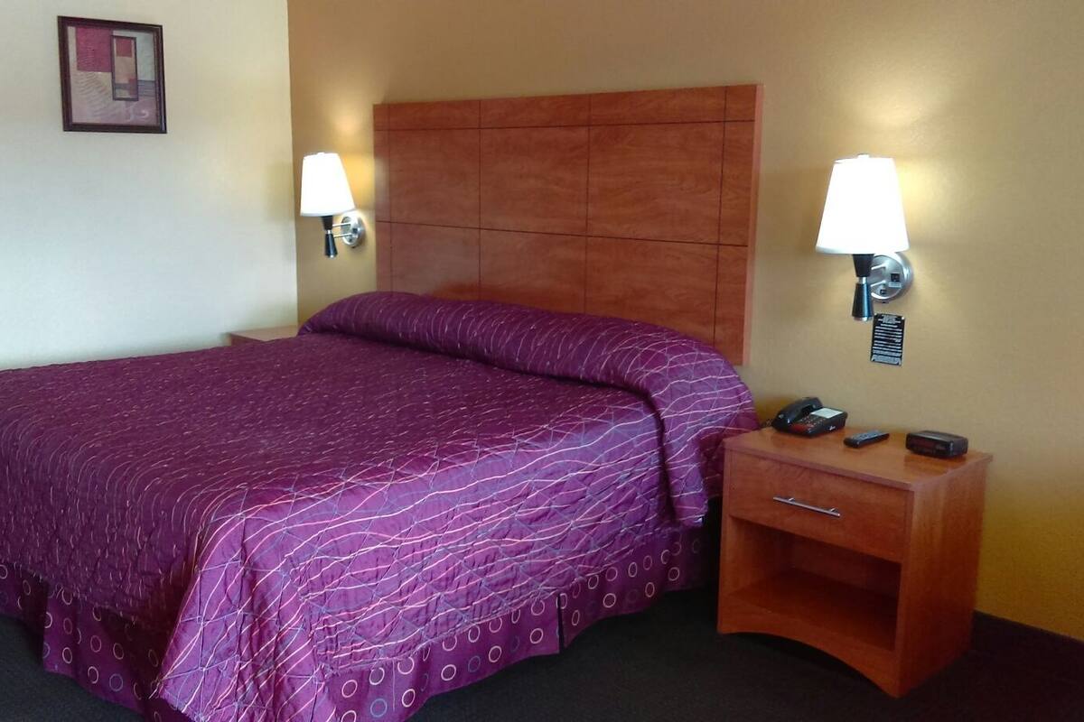 Une chambre d’hôtel avec un lit, deux lampes de chevet, une table de chevet avec un téléphone et des télécommandes, et un tableau encadré au mur.