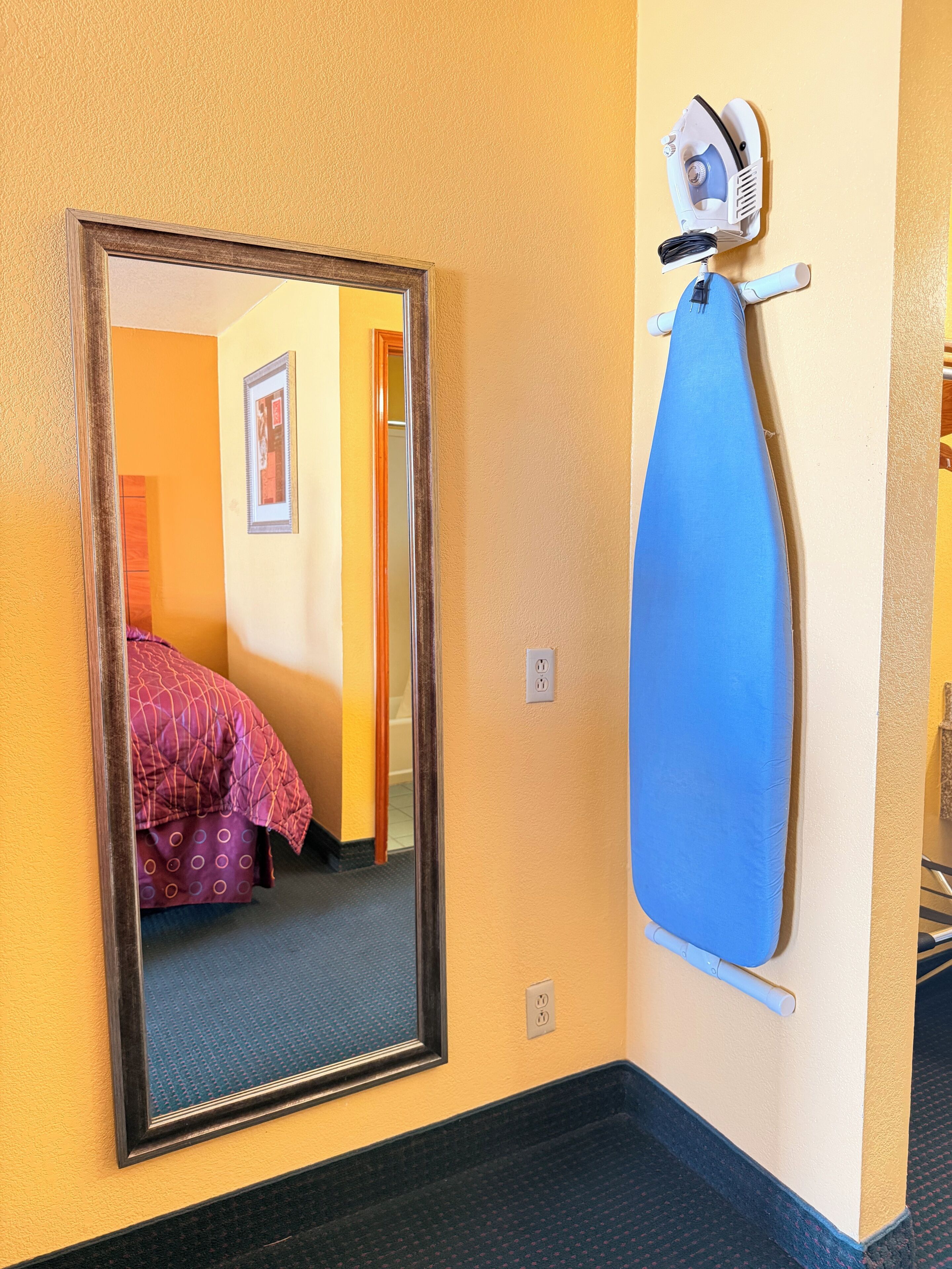 A room with a large mirror, a blue ironing board, and a bed with a patterned blanket.