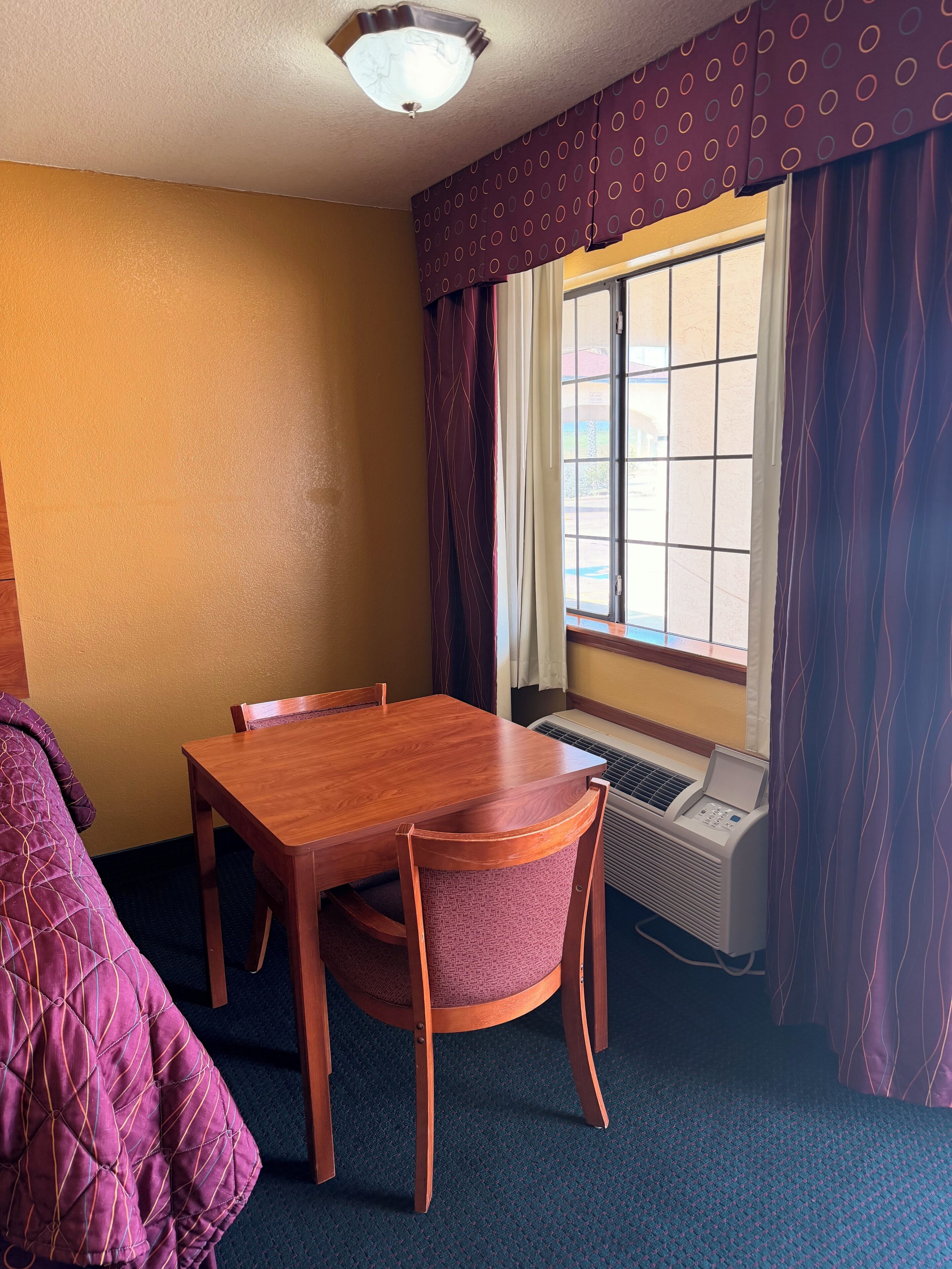 Una habitación con una mesa de madera, sillas, un ventanal con cortinas y un equipo de aire acondicionado.