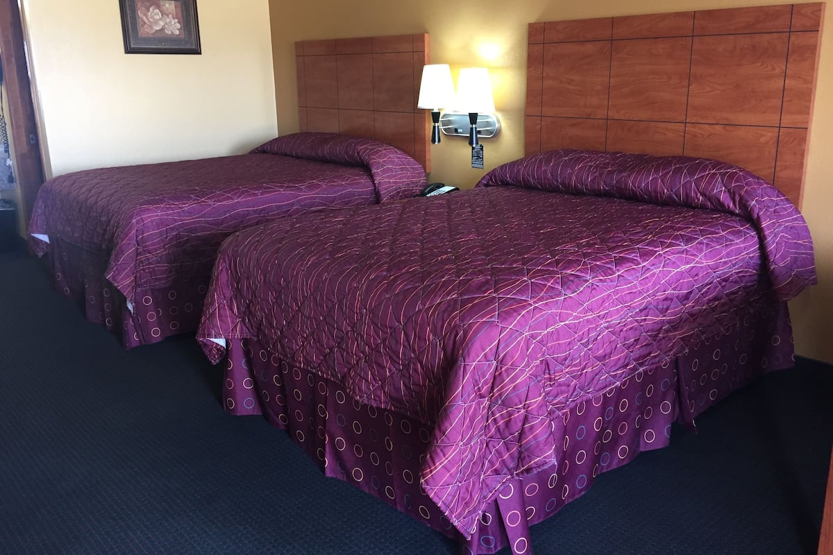 Une chambre d’hôtel avec deux lits, chacun recouvert d’une couvre-lit violette et agrémenté d’oreillers assortis. Une lampe est fixée au mur entre les deux lits, et un tableau encadré est accroché au mur.