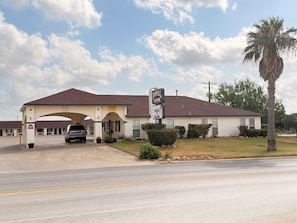 A motel with a single-story structure, a covered parking area, and a palm tree in the foreground. - Royal Inn Pearsall (Pearsall)