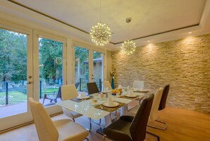 A modern dining room with a glass table, stone wall, and large windows overlooking a garden. - Riverfront Bed and Breakfast (District of North Vancouver)