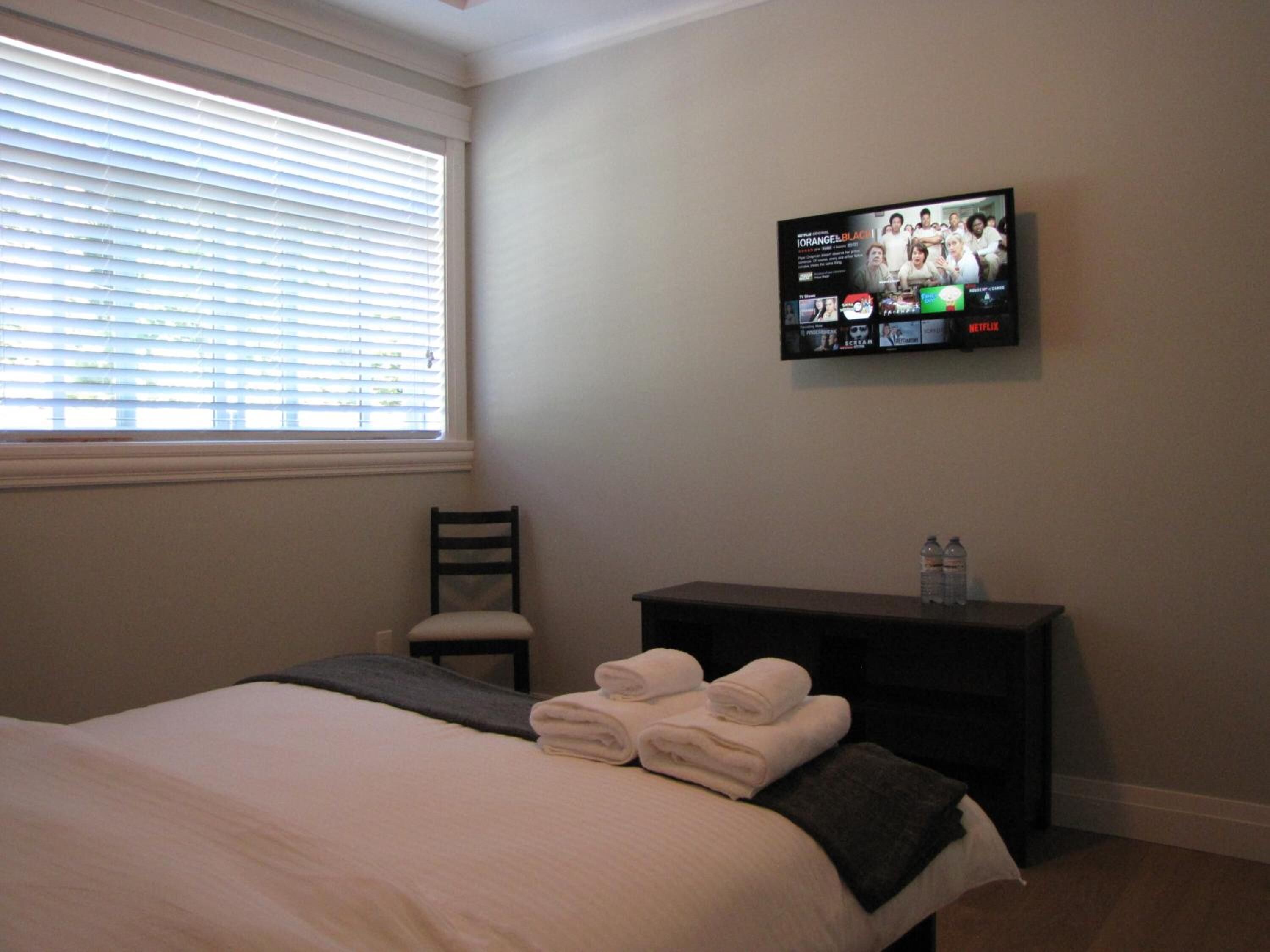 A hotel room with a bed, a chair, a TV mounted on the wall, and a small table with towels and a water bottle.