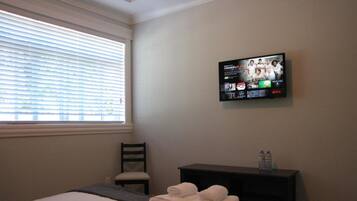 A hotel room with a bed, a chair, a TV mounted on the wall, and a small table with towels and a water bottle.