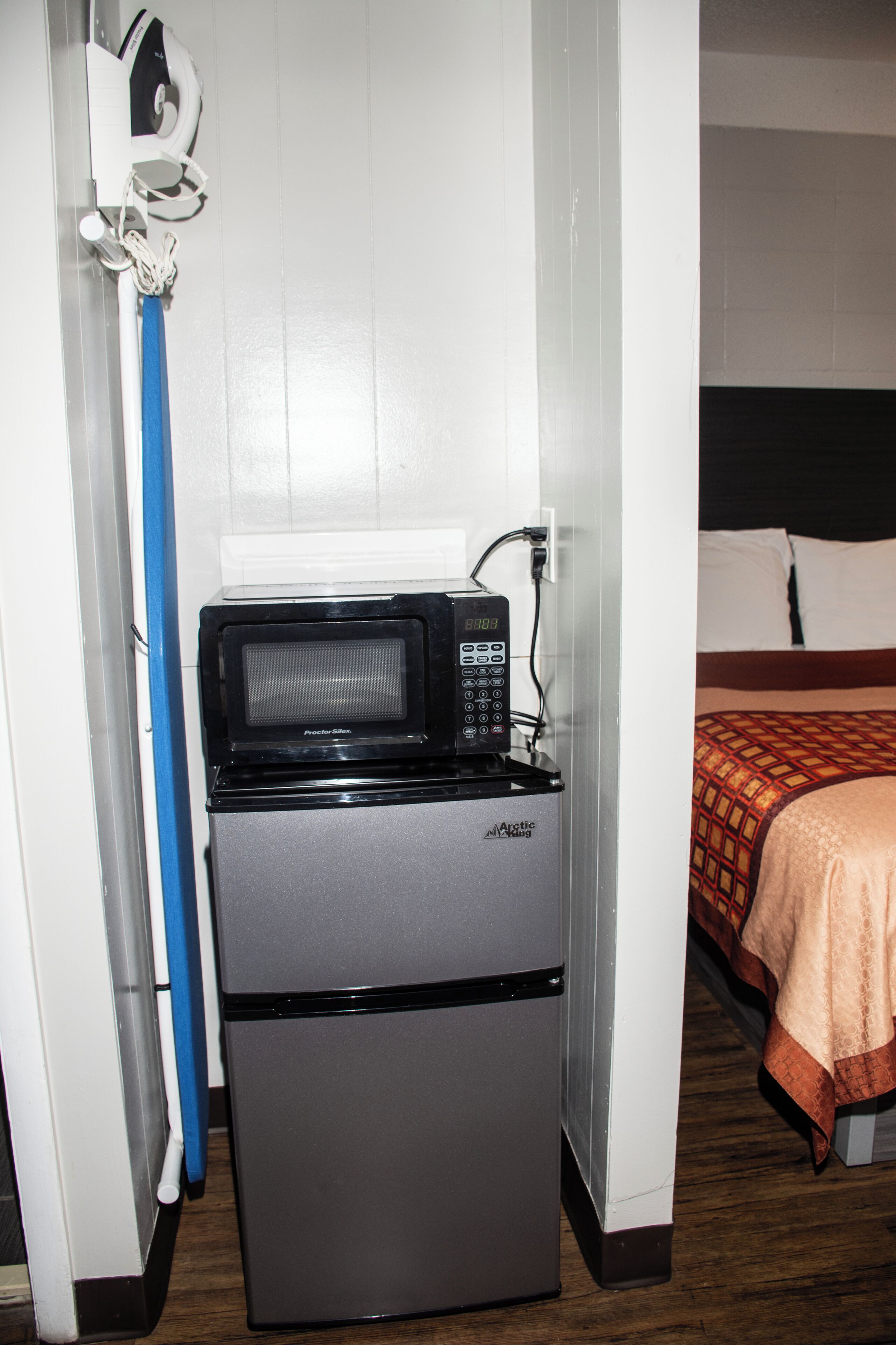 A compact kitchenette with a microwave and refrigerator, a blue cleaning tool, and a bed with a patterned blanket.