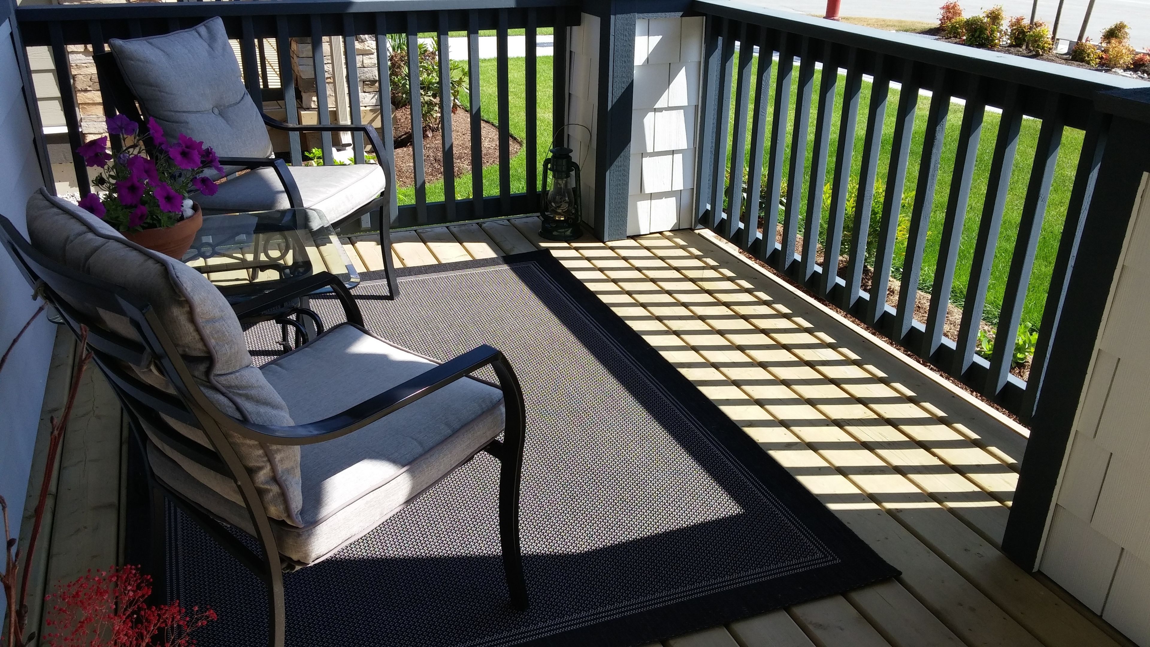 A covered outdoor seating area with two chairs and a table, a black mat, and a view of a garden.