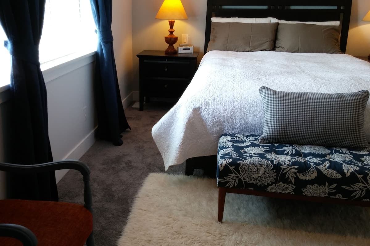 Une chambre à coucher comprenant un lit, une table de chevet, une lampe, une chaise et une fenêtre avec des rideaux.
