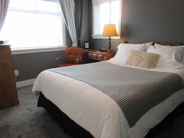 A neatly made bed with a white comforter and a patterned throw, a wooden nightstand with a lamp, a chair, and a desk with a computer. - Seagrass Guesthouse (Tsawwassen)