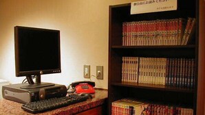 A computer desk with a monitor, keyboard, and telephone. A bookshelf with books is visible in the background. - Hotel Takamatsu Hills (Takamatsu)