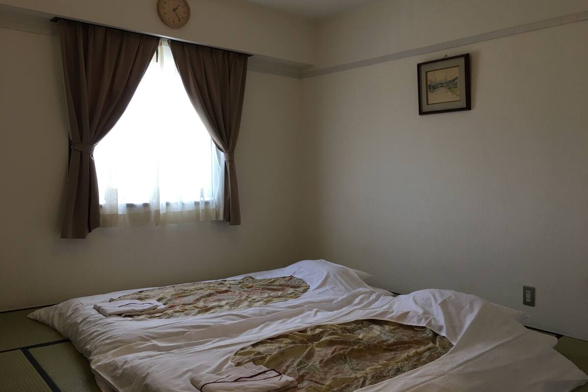 Ein Zimmer mit einem Bett, einem Vorhangfenster, einer Uhr und einem gerahmten Bild an der Wand.