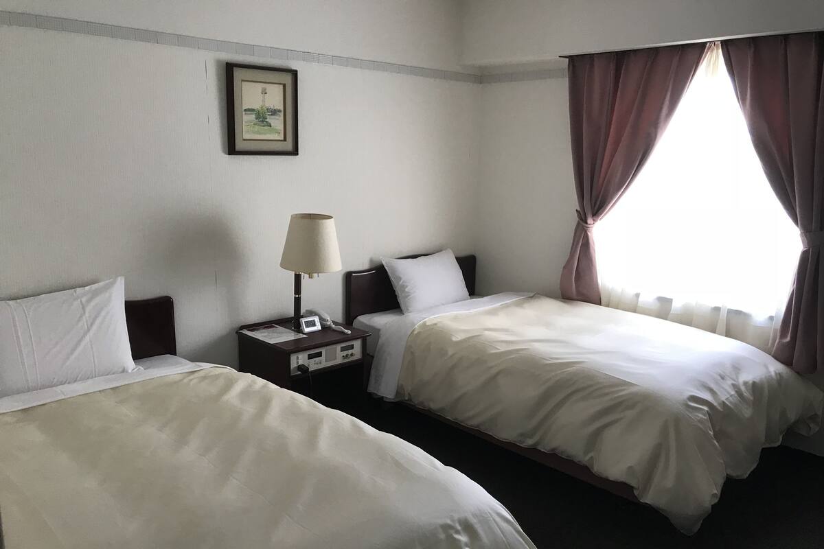 Ein Hotelzimmer mit zwei Betten, einem Nachttisch mit Lampe, einem gerahmten Bild an der Wand und einem Fenster mit Vorhängen.