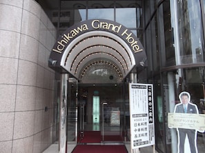 De ingang van het Ichikawa Grand Hotel met een glazen deur, een rode loper en een bord met informatie over het hotel.