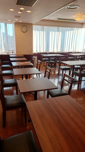 Un comedor amplio con varias mesas y sillas de madera, ventanas grandes con cortinas translúcidas y un reloj en la pared.