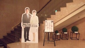 A lobby area with a marble staircase, a poster of a man and woman, and a stand with a schedule or list. - Ichikawa Grand Hotel (Ichikawa)