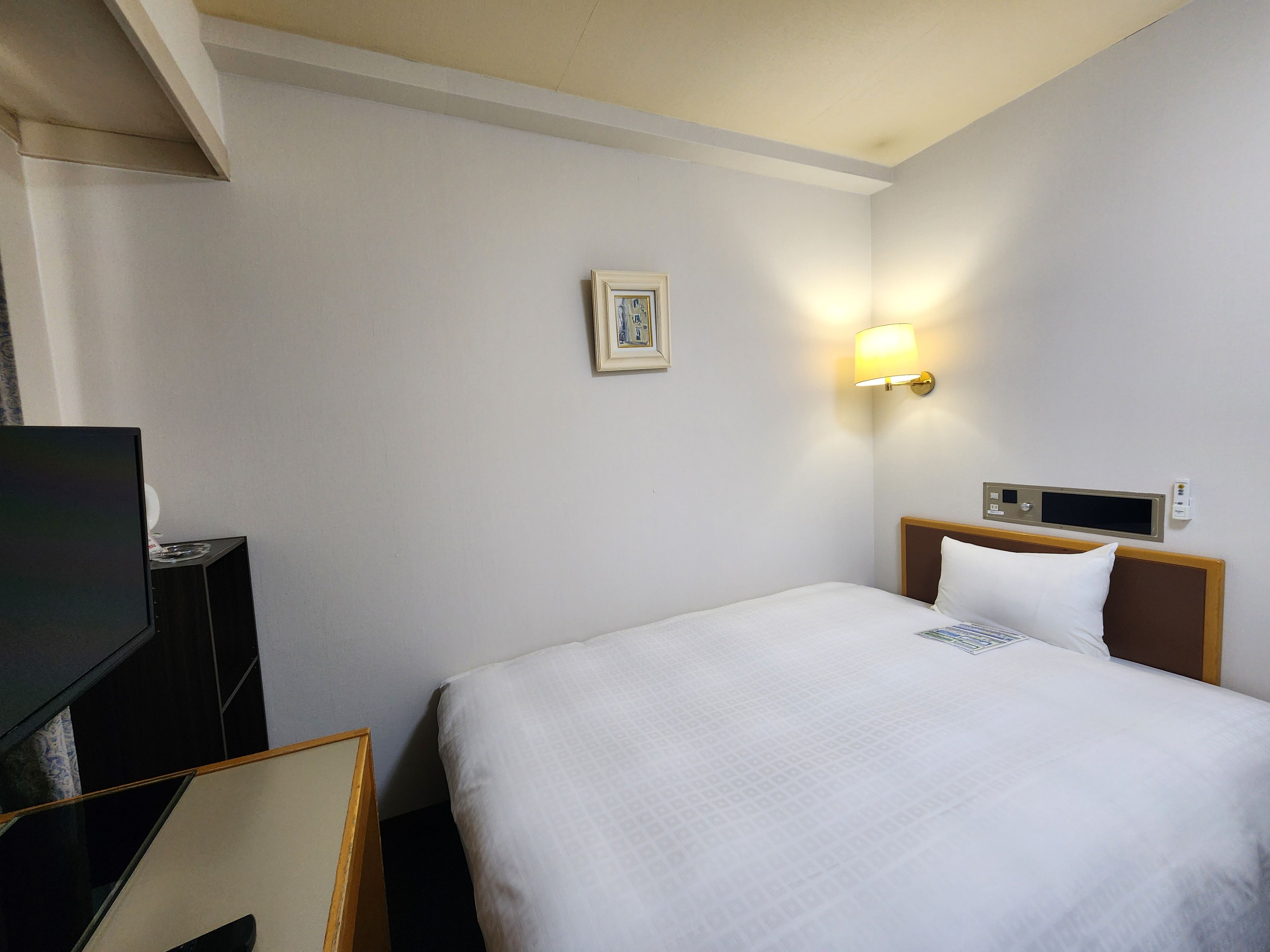 A hotel room with a bed, a bedside table, a television, and a wall-mounted light.