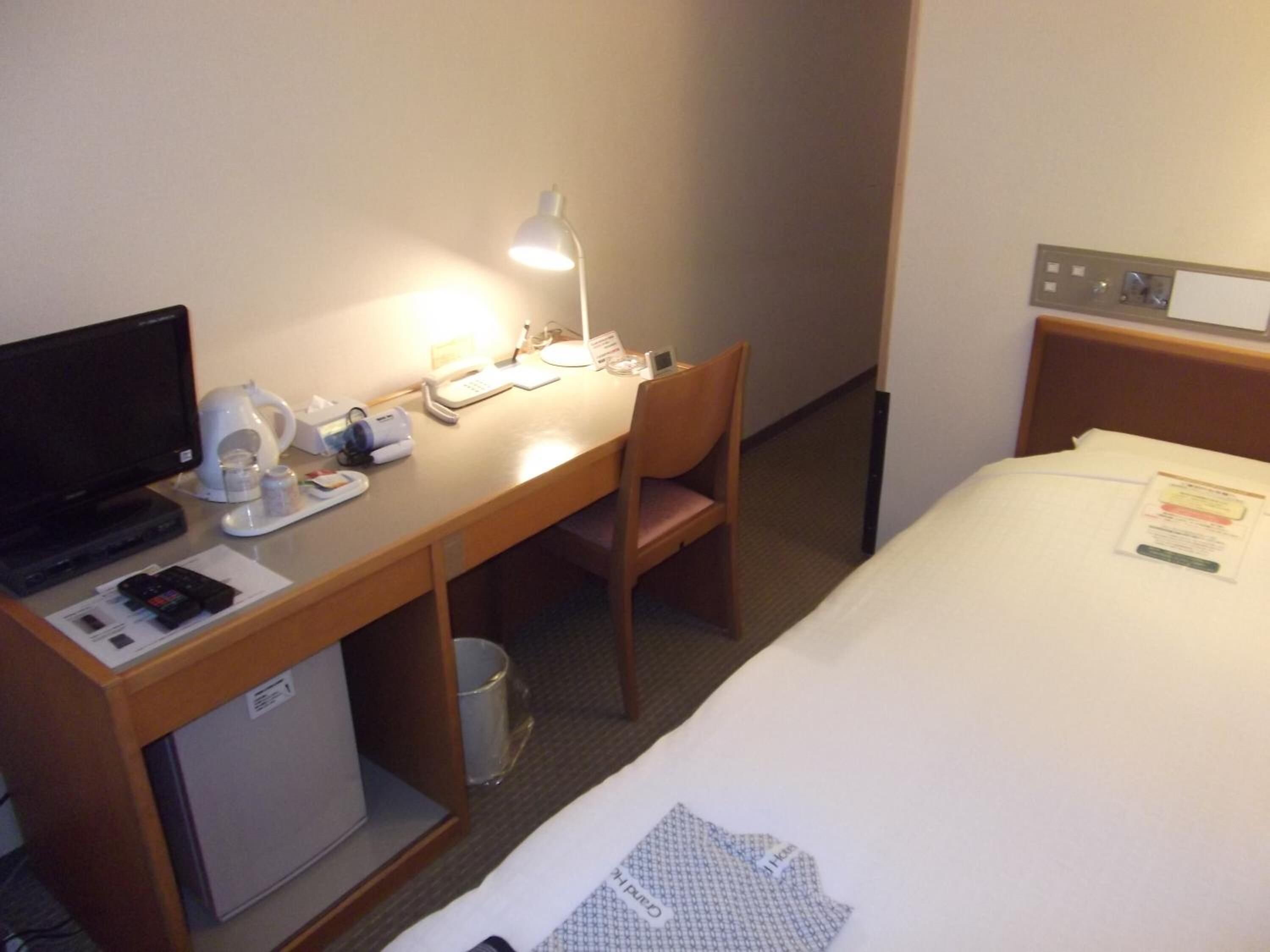 Ett hotellrum med en säng, ett skrivbord med en dator, en lampa, en stol och ett litet kylskåp.