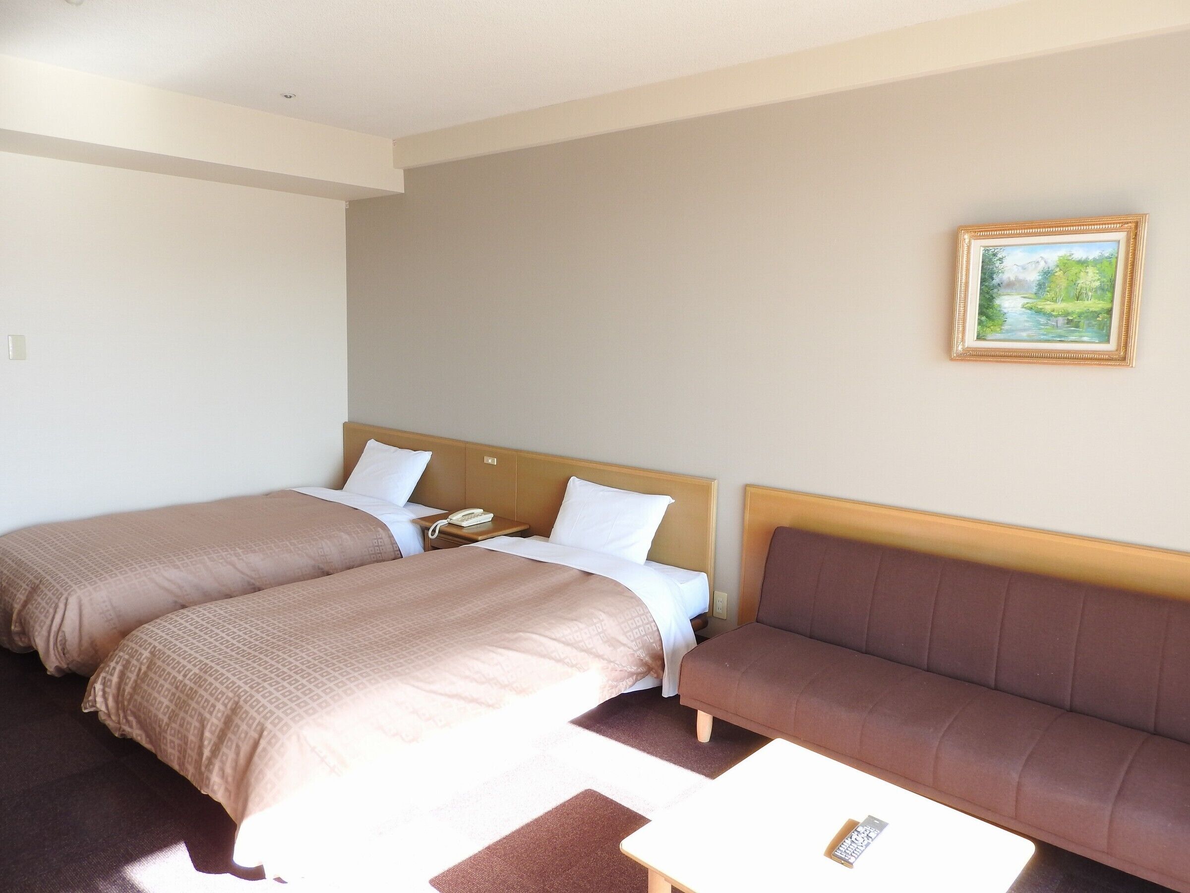 A hotel room with two beds, a sofa, a painting on the wall, and a remote control on the sofa.