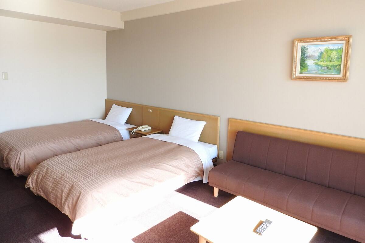A hotel room with two beds, a sofa, a painting on the wall, and a remote control on the sofa.