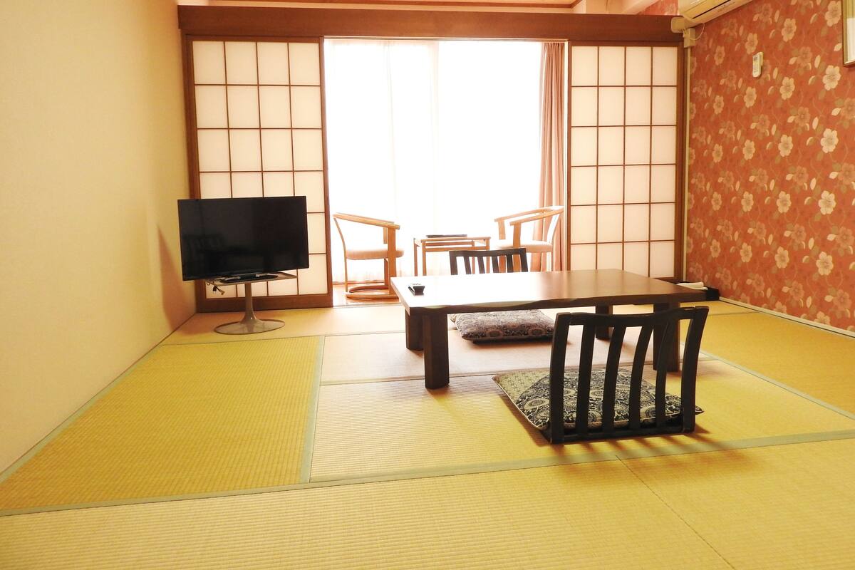 A traditional Japanese room with tatami flooring, a low table, chairs, a television, and sliding doors.
