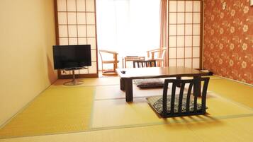 Una habitación tradicional japonesa con suelo de tatami, una mesa baja, sillas, un televisor y puertas correderas.