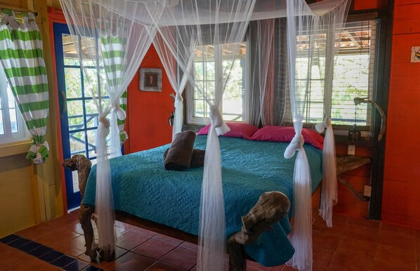 Una habitación con cama bajo un dosel, una ventana con cortinas y una mesita de noche con lámpara.