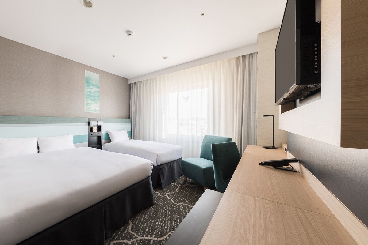 Une chambre d’hôtel avec deux lits, un bureau, une chaise, une télévision et une fenêtre avec des rideaux.