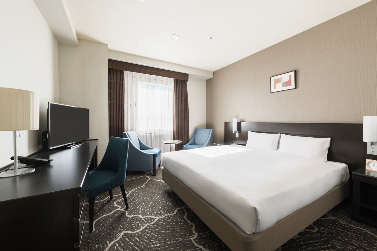 Une chambre d’hôtel avec un grand lit, un bureau avec une télévision, deux chaises, une petite table et une fenêtre avec des rideaux.
