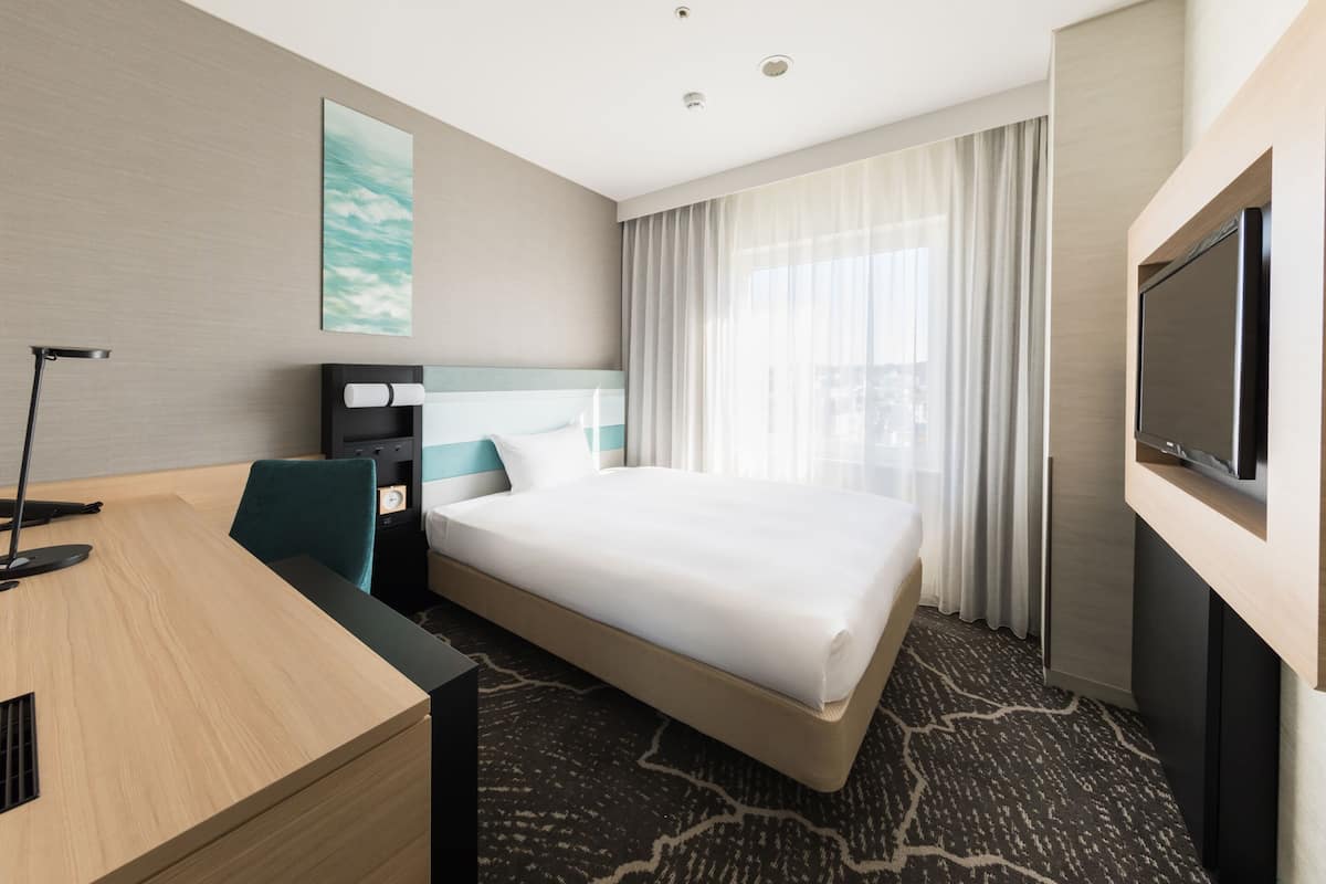 Ein modernes Hotelzimmer mit einem großen Bett, einem Schreibtisch, einem Stuhl, einem Flachbildfernseher und einem Fenster mit Vorhängen.