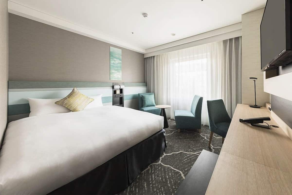 Ein Hotelzimmer mit einem großen Bett, einem Schreibtisch mit Fernseher, einem Sessel, einem kleinen Tisch und einem Fenster mit Vorhängen.